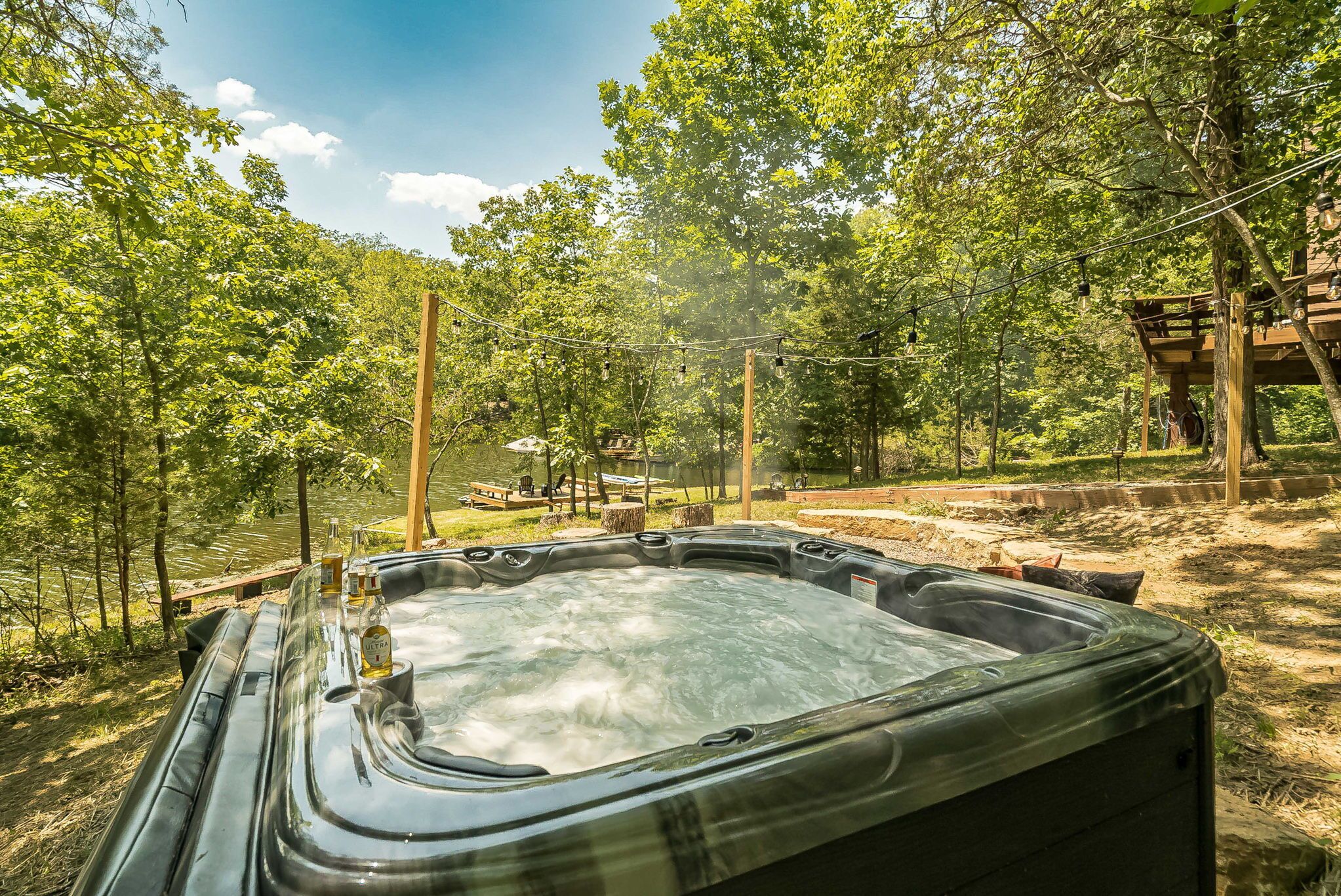 Un bain à remous situé dans un espace boisé, avec une terrasse en bois et des tables de pique-nique en arrière-plan.
