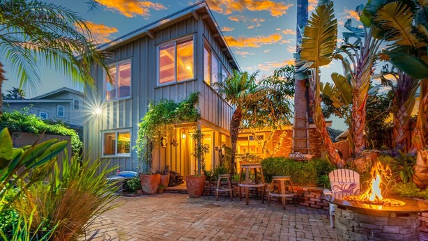 A two-story house with a patio area featuring a fire pit, outdoor furniture, and lush tropical plants.