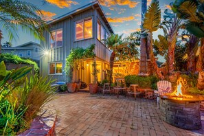 Una casa de dos pisos con un patio que cuenta con una fogata, mobiliario exterior y exuberante vegetación tropical.
