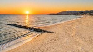 A beach with a pier extending into the water, a sunset in the background, and a distant mountain range.