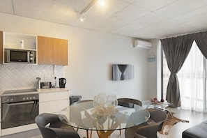 A modern kitchen with a microwave, oven, and coffee maker, a glass dining table with chairs, and a living area with a TV and curtains.