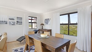 Ein Essbereich mit einem Holztisch, beigen Stühlen und einem Teppich. An der Wand hängen gerahmte Bilder und große Fenster bieten einen Blick ins Freie.