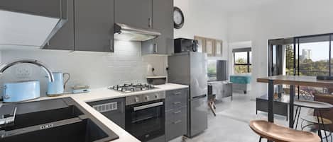 Una cocina moderna con armarios grises, un refrigerador de acero inoxidable, una estufa a gas y un fregadero. Se ve un área de comedor con una mesa y sillas a través de una puerta abierta.
