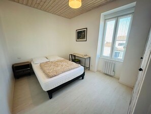 A bedroom with a bed, bedside table, chair, window, radiator, and a picture on the wall. - Cocon St Hilaire, charming house, enclosed garden (Talmont-Saint-Hilaire)