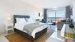 Quarto de hotel moderno com uma cama grande, área de estar com sofá e poltronas, televisão e vista para a cidade.