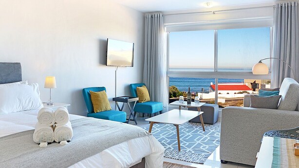 Quarto de hotel com cama, duas poltronas azuis, um sofá, uma mesa pequena e vista para o mar.