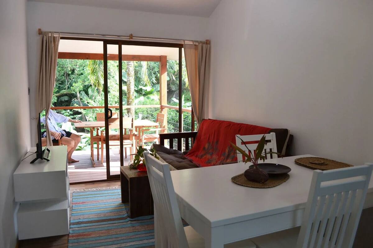 Une pièce comprenant une table à manger, des chaises, un canapé, un téléviseur et donnant sur l’extérieur.