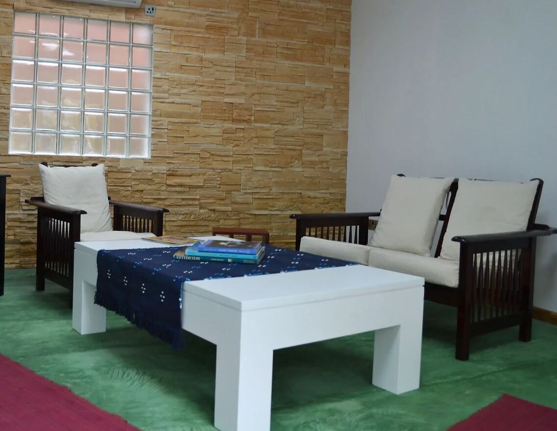A room with a stone wall, a window with blinds, a white sofa, two armchairs, a white coffee table, and a green carpet.