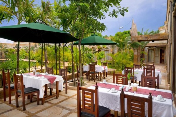 An outdoor dining area with tables and chairs, surrounded by greenery and a stone structure.