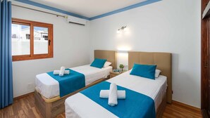 Une chambre d’hôtel avec deux lits, chacun recouvert d’une couvre-lit bleue et doté de draps blancs, avec une tête de lit en bois, une fenêtre avec des rideaux et un luminaire fixé au mur.
