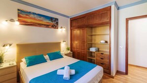 Un lit bien fait, avec une couverture bleue et des oreillers, une armoire en bois et un tableau représentant un coucher de soleil accroché au mur.