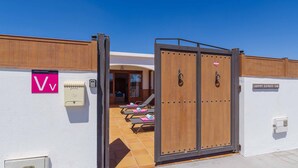 L’entrée d’un hôtel, avec une porte en bois, une enseigne indiquant une chambre de catégorie « V+ », et un espace piscine équipé de chaises longues.