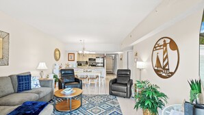 Een moderne woonkamer met een bank, fauteuils, een salontafel en een kamerkleed. Achtergrond toont een keuken met witte kasten en een eethoek.
