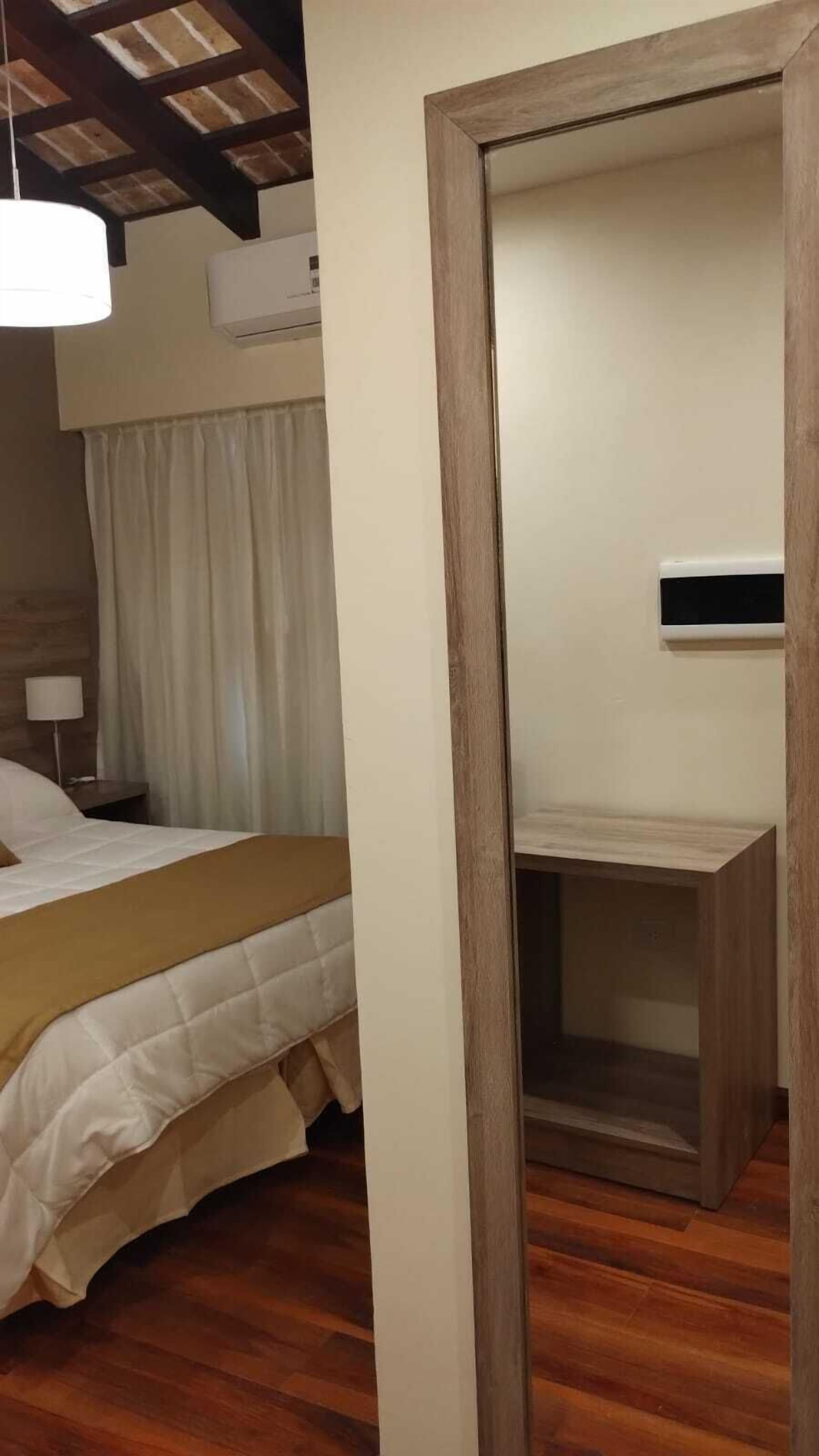 Una habitación de hotel con piso de madera, cama con ropa blanca, mesita de noche, lámpara y ventilador de techo.