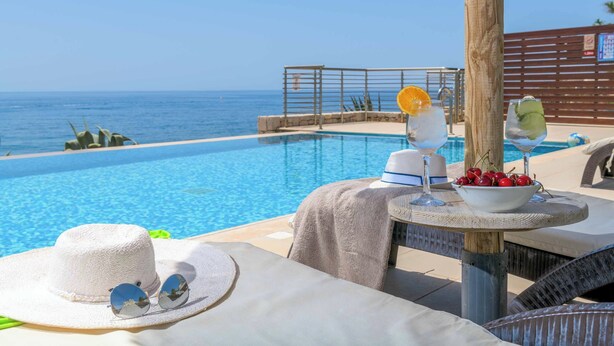 Zona junto a la piscina con vista al mar, una mesa con un bol de cerezas, un sombrero y gafas de sol.