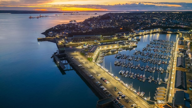 An aerial view of a marina with boats docked, a road leading to the marina, and a sunset in the background. - Fulke Street - 2 Bedroom Apartment - Milford Haven (Milford Haven)