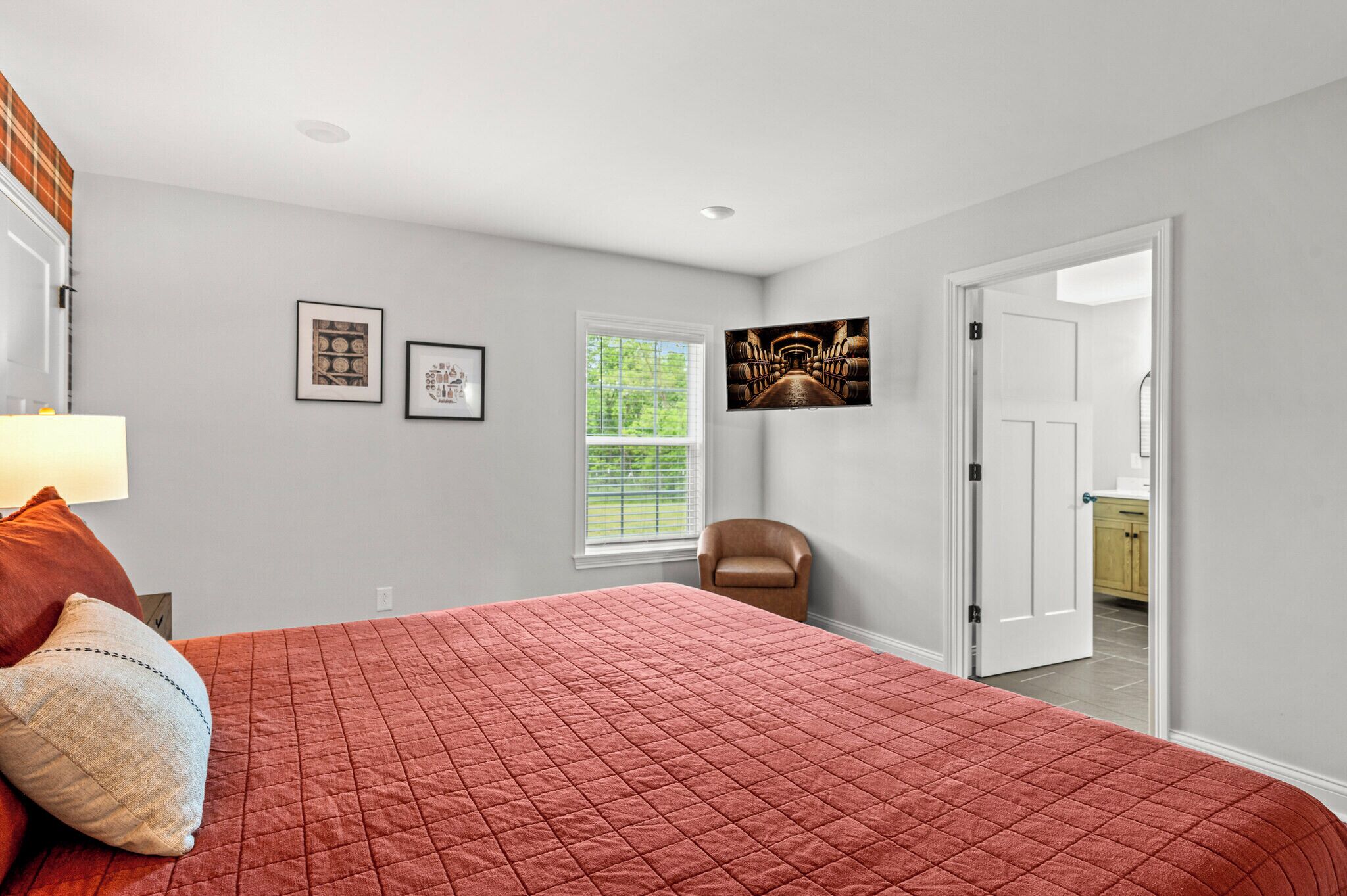 A bedroom with a red quilted bedspread, a chair, a window with blinds, and framed pictures on the wall.