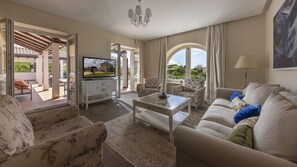 A living room with a sofa, armchairs, a coffee table, and a TV. There is a view of a garden through large windows.