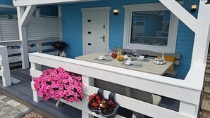 A covered outdoor seating area with a table set for two, a white door, and a window with blinds.