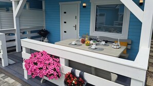 Ein überdachter Außenbereich mit Tisch für zwei Personen, einer Bank mit rosafarbenen Blumen und einer Tür rechts.