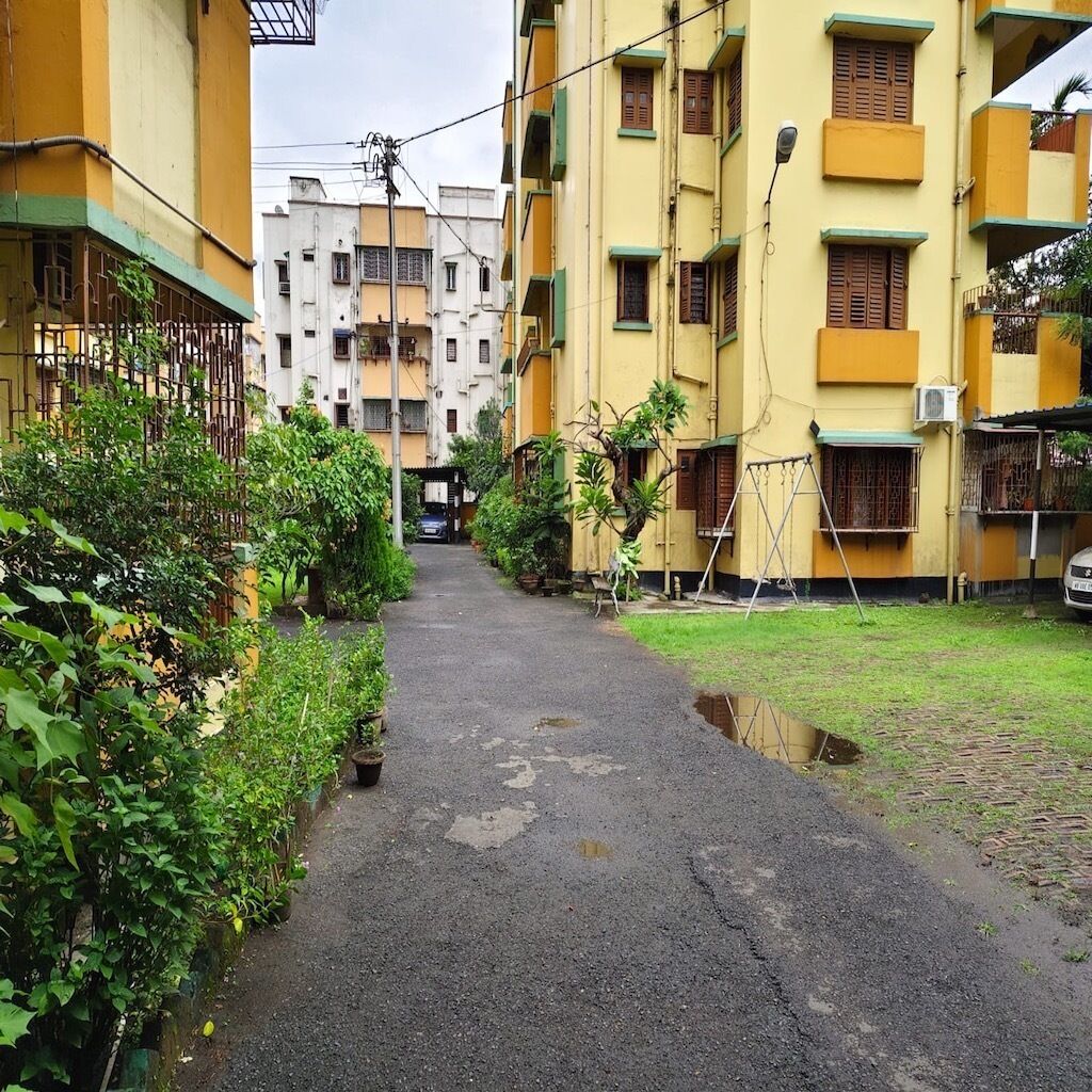 Uma passagem estreita entre edifícios residenciais, com um caminho pavimentado, plantas em vasos e uma pequena área gramada.