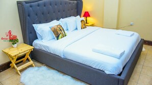 Una cama bien hecha con cabecera gris, ropa de cama blanca y almohada con estampado de elefante. Hay una mesita de noche de madera con una lámpara y un florero.