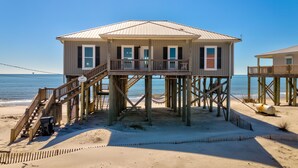 A wooden beach house with a balcony, a staircase, and a sandy beach in the background.