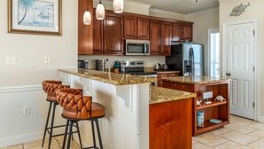 A kitchen with wooden cabinets, a granite countertop, a stainless steel microwave, and a refrigerator. There are three bar stools with leather seats.