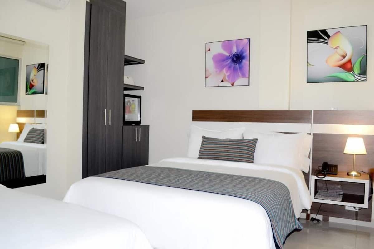 Hotellihuone, jossa on suuri sänky, yöpöytä lampun kanssa ja taideteoksia seinillä.