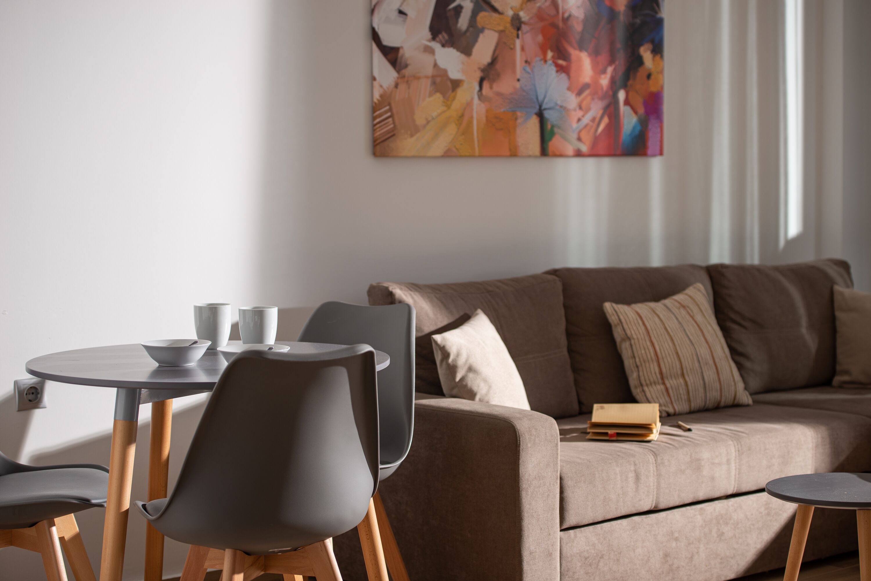 A modern living room with a beige sofa, a round dining table, and two chairs. A colorful abstract painting is on the wall.