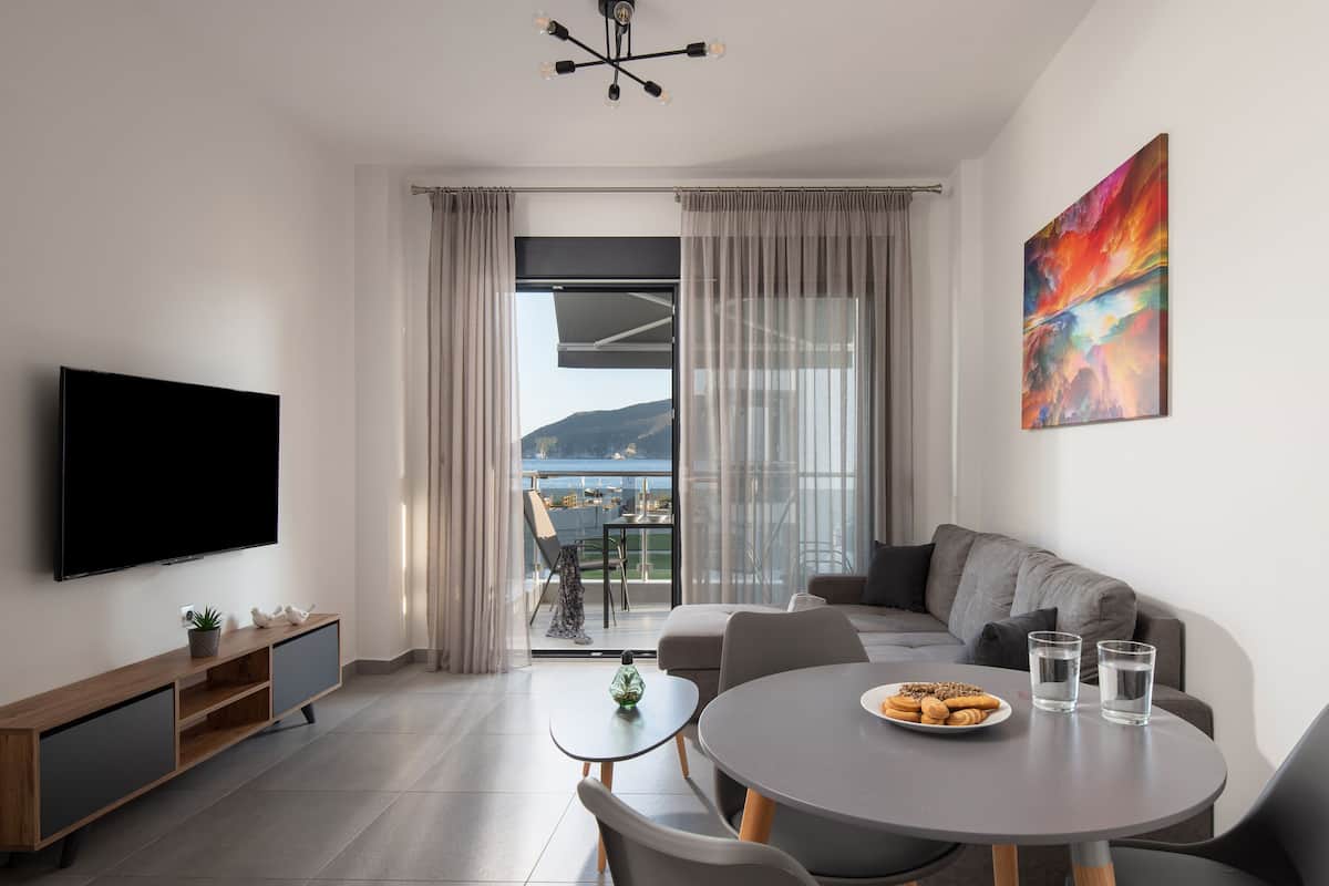 A modern living room with a flat-screen TV, a round dining table with two chairs, a sofa, and a balcony with a view of the sea.