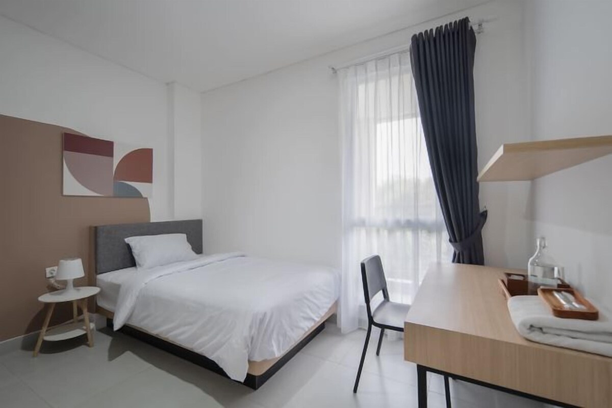 A modern hotel room with a bed, a desk, a chair, and a window with sheer curtains.