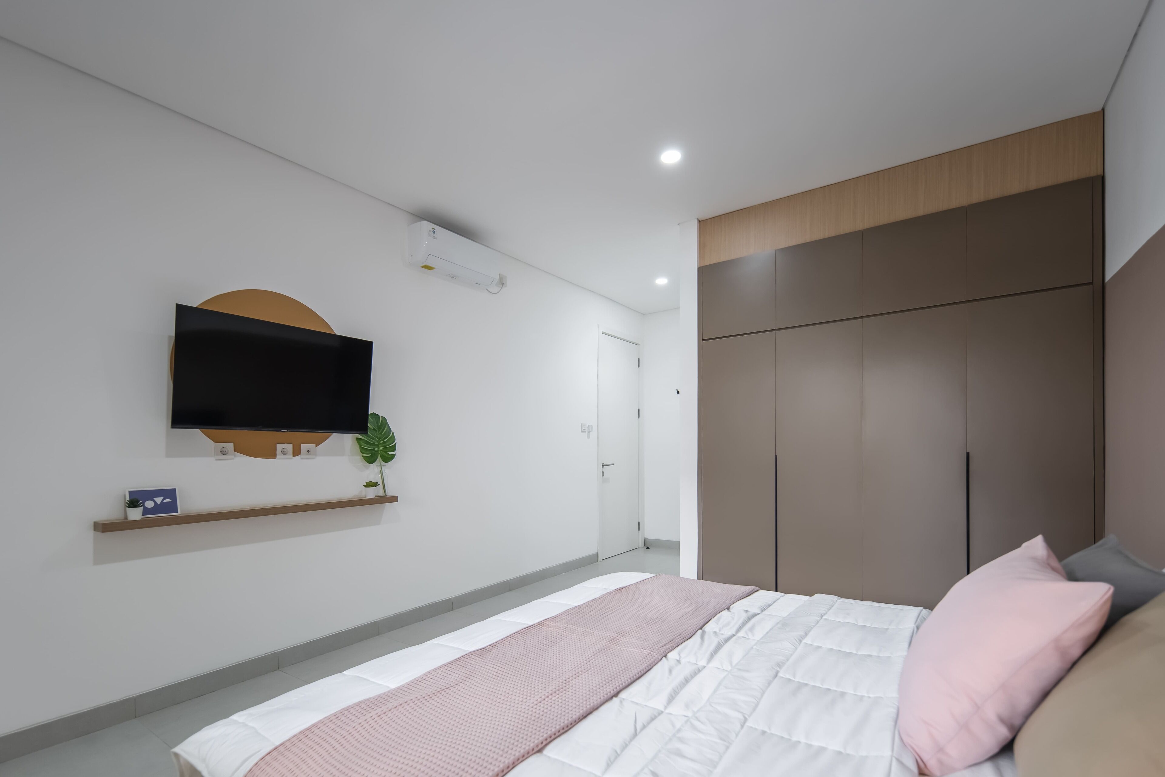 A modern bedroom with a bed, a TV mounted on the wall, a wardrobe, and a small shelf with a plant.
