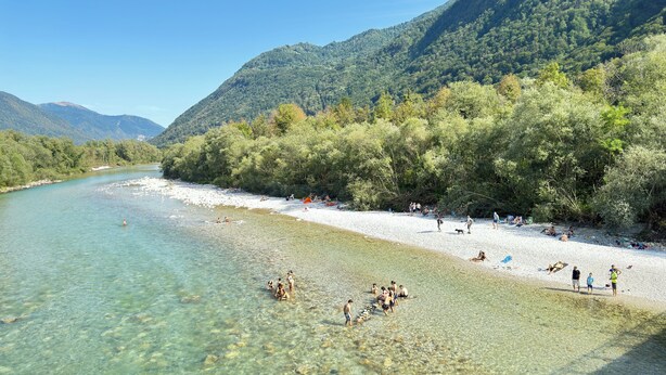 A riverbank with clear water, people relaxing, and a forested hillside in the background.