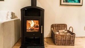 A modern wood-burning stove with a glass front, a wicker basket filled with firewood, and a framed painting on the wall.