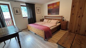 Quarto com cama de madeira, mesa de cabeceira, uma mesa e uma janela com cortinas.