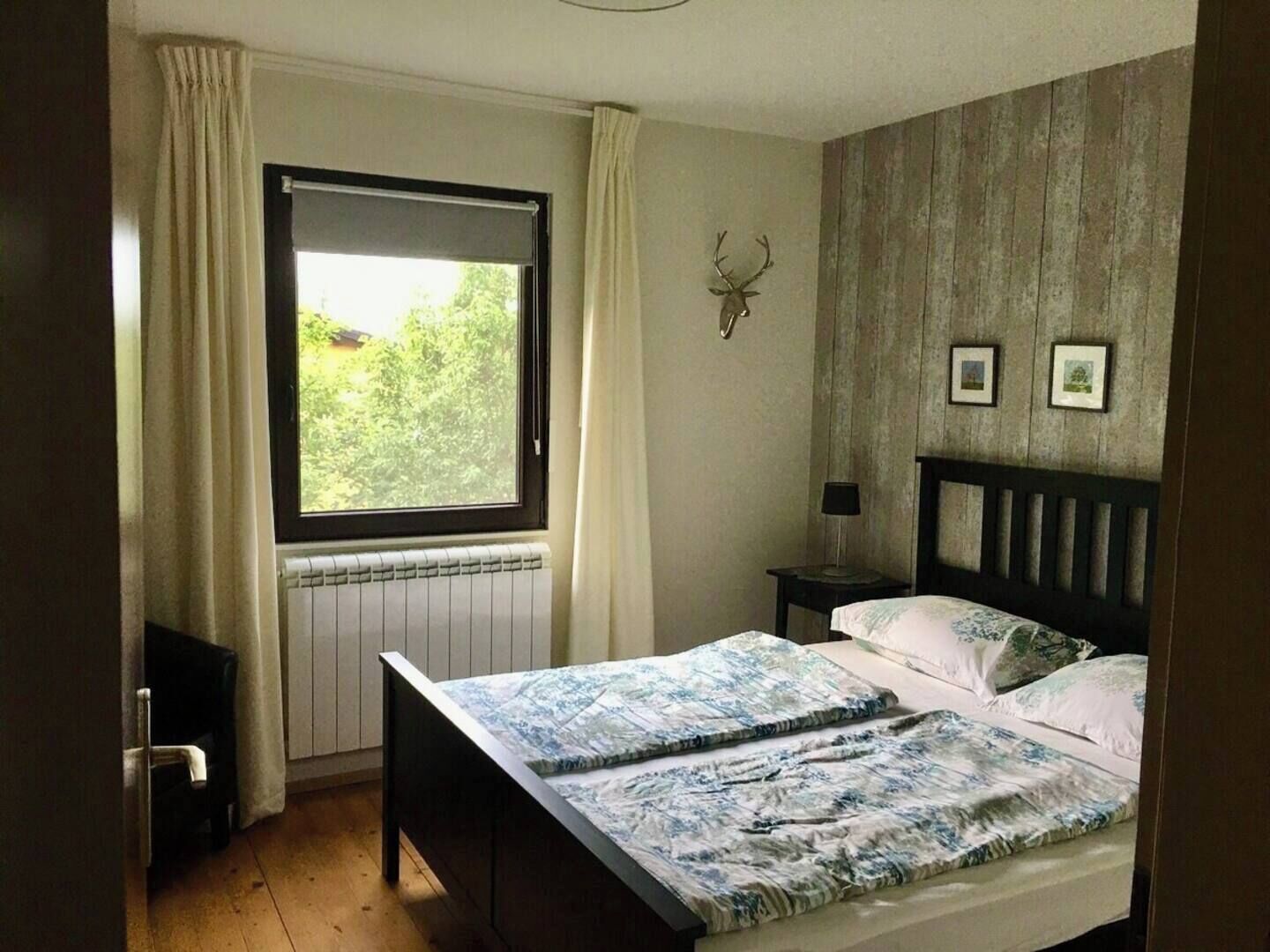 Un dormitorio con cama, ventana, radiador, mesita de noche, lámpara y un adorno de cabeza de ciervo montado en la pared.