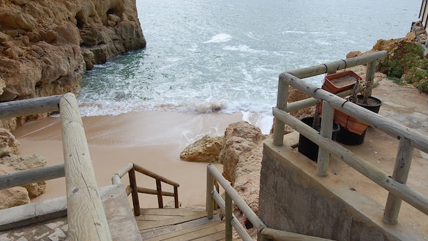 Un escalier en bois mène à une plage bordée d’une falaise rocheuse et dotée d’une rambarde en bois.