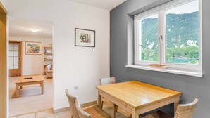 A dining area with a wooden table and chairs, a window with a view of a forested area, and a shelf with decorative items.