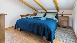 A bedroom with a sloped ceiling, wooden beams, a bed with blue bedding, two nightstands, and a patterned rug.