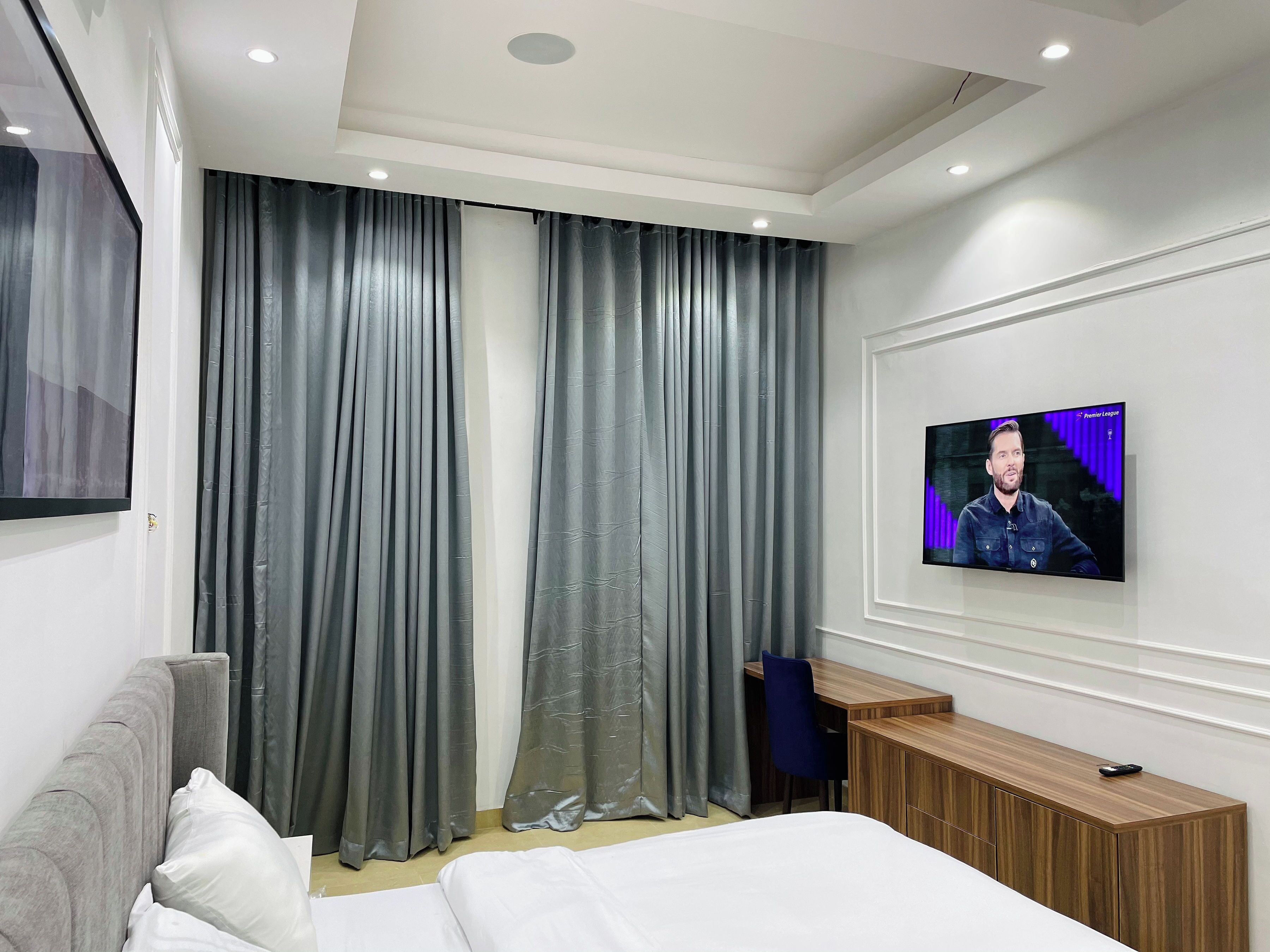 A modern hotel room with a large bed, a desk, a TV mounted on the wall, and grey curtains.