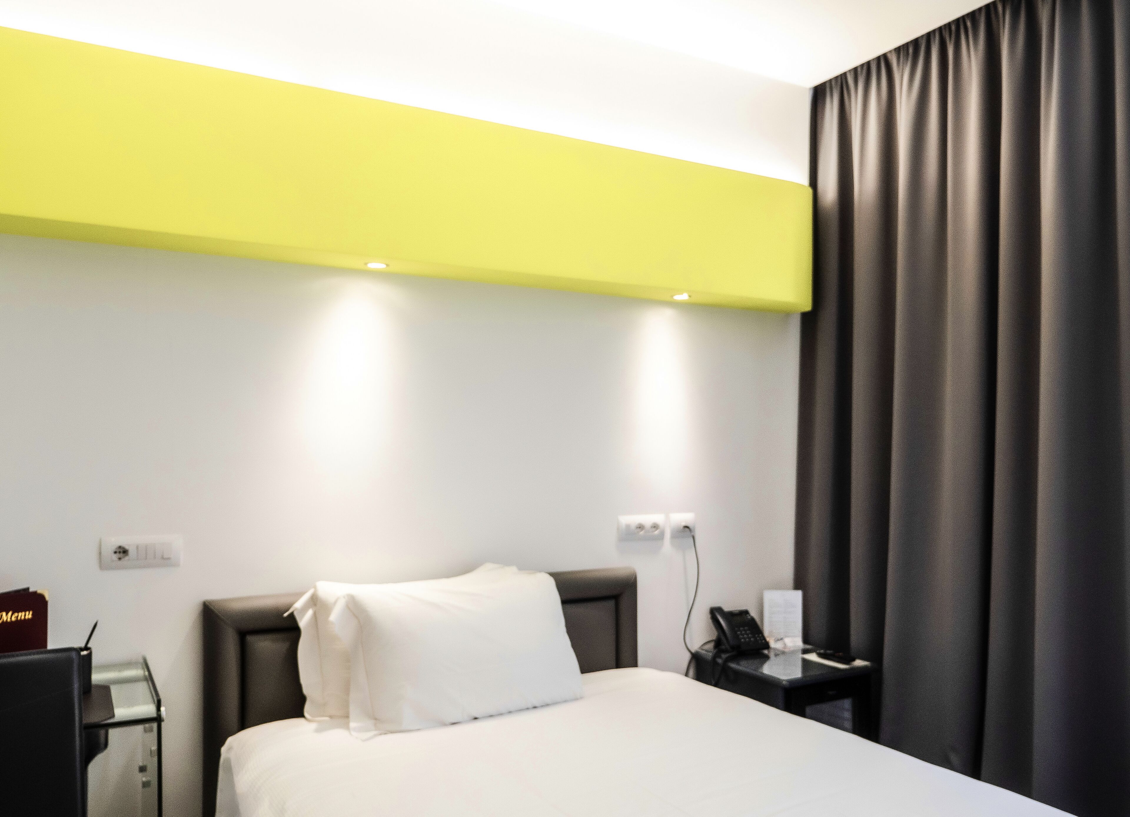 A hotel room with a bed, a bedside table with a phone and a menu, and a wall socket.