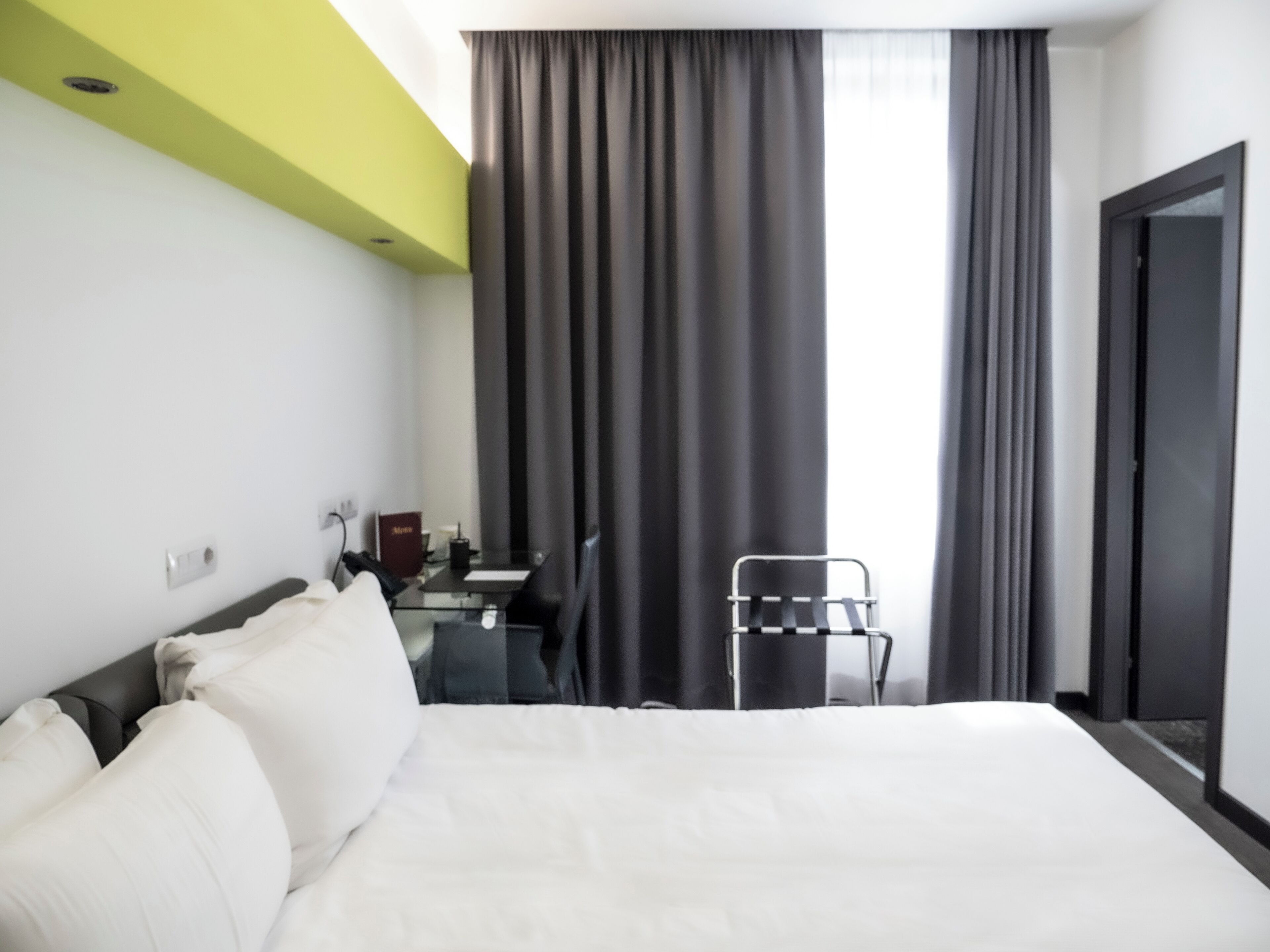 A hotel room with a large bed, a desk with a chair, a window with curtains, and a door.