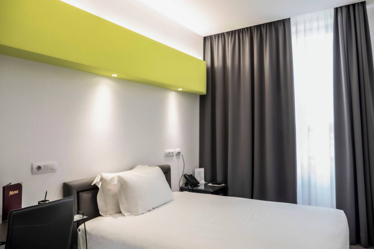 A modern hotel room with a large bed, a desk, and a chair. There is a window with curtains, a wall-mounted light, and a power outlet.