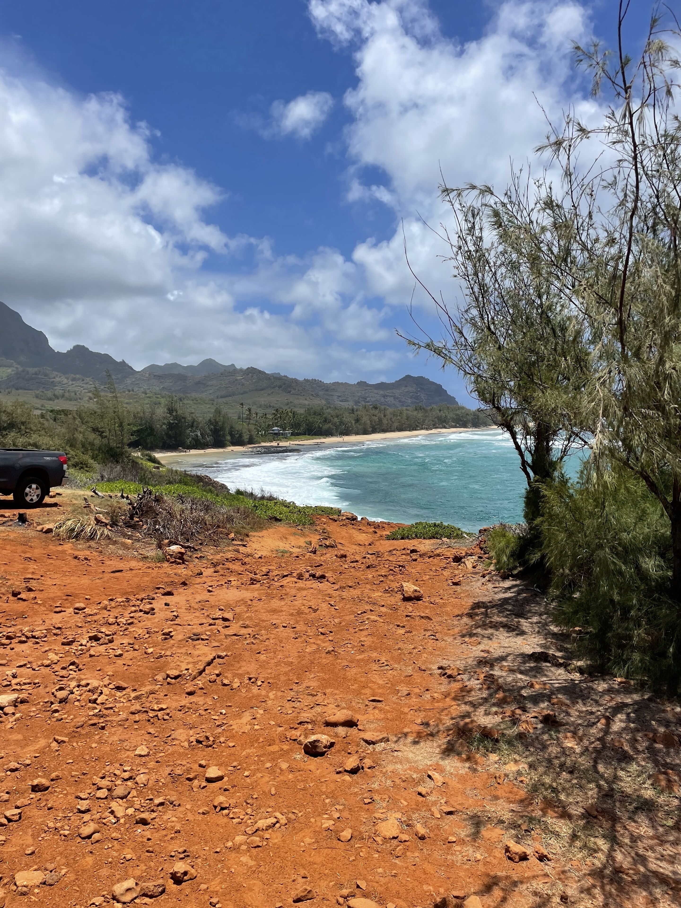 A coastal landscape with a dirt path leading to a beach, a parked vehicle, and a mountain in the background.