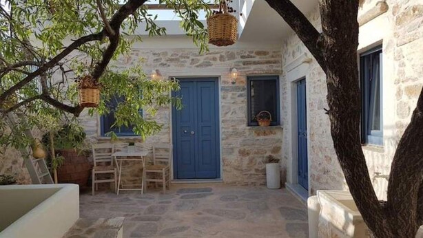 A stone building with a blue door, a small table with chairs, and hanging baskets. - 1875 panoramic stone house (Filoti)
