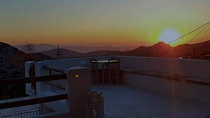 A sunset view from a balcony with a railing, a chair, and a small table. - 1875 panoramic stone house (Filoti)