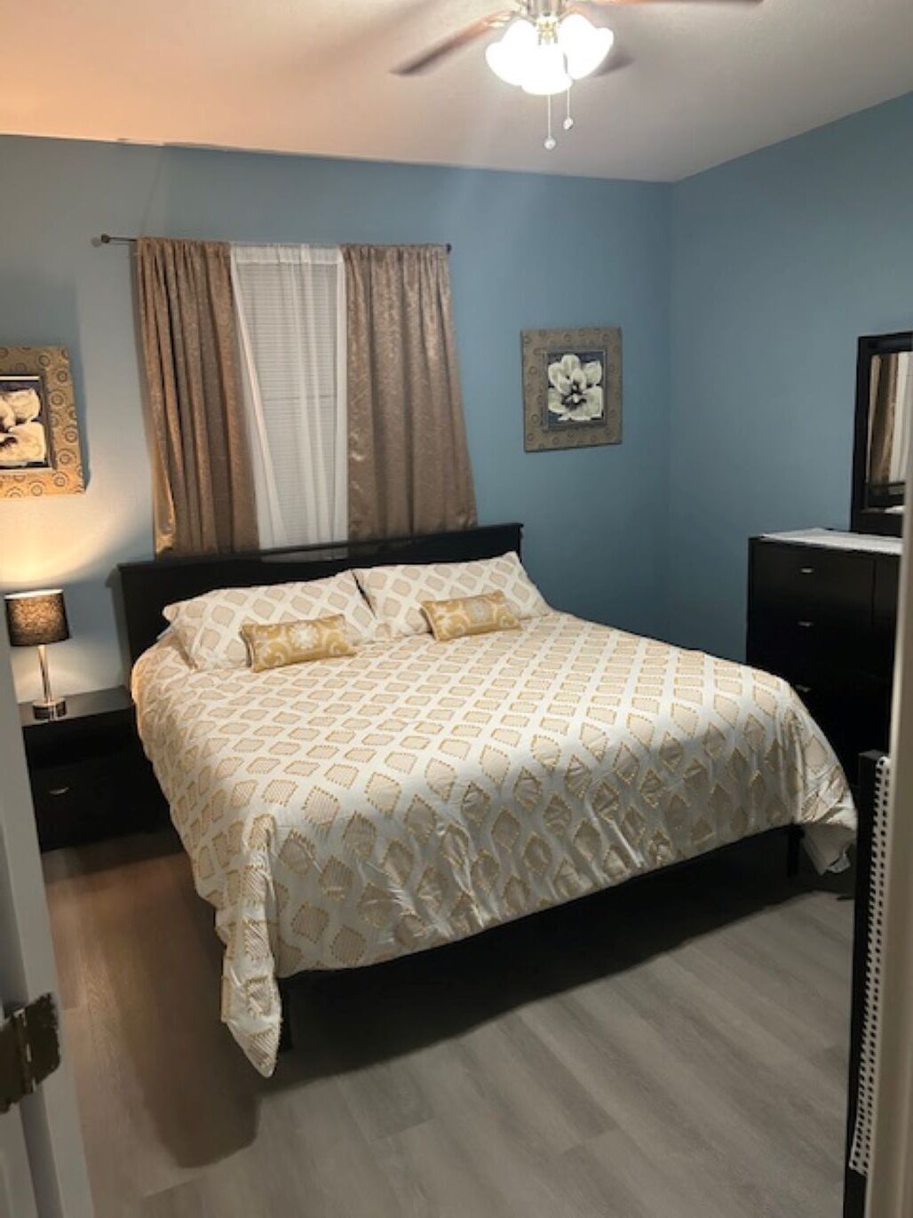 Quarto com cama, mesa de cabeceira, cômoda, ventoinha no teto e um quadro pendurado na parede.