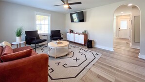 A modern living room with a ceiling fan, a TV mounted on the wall, a round coffee table, and a patterned rug. - 2 BR King Bed Near Duke Hospital & Campus (Durham)