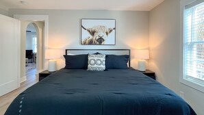 A bedroom with a large bed, two bedside lamps, a framed artwork of a bull, and a window with blinds. - 2 BR King Bed Near Duke Hospital & Campus (Durham)